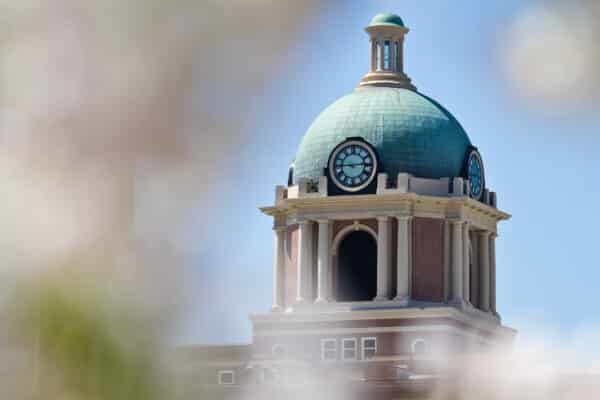 A classical domed clock tower with a teal roof and three clock faces against a clear blue sky. The brick structure features neoclassical architectural details, with a soft foreground blur.