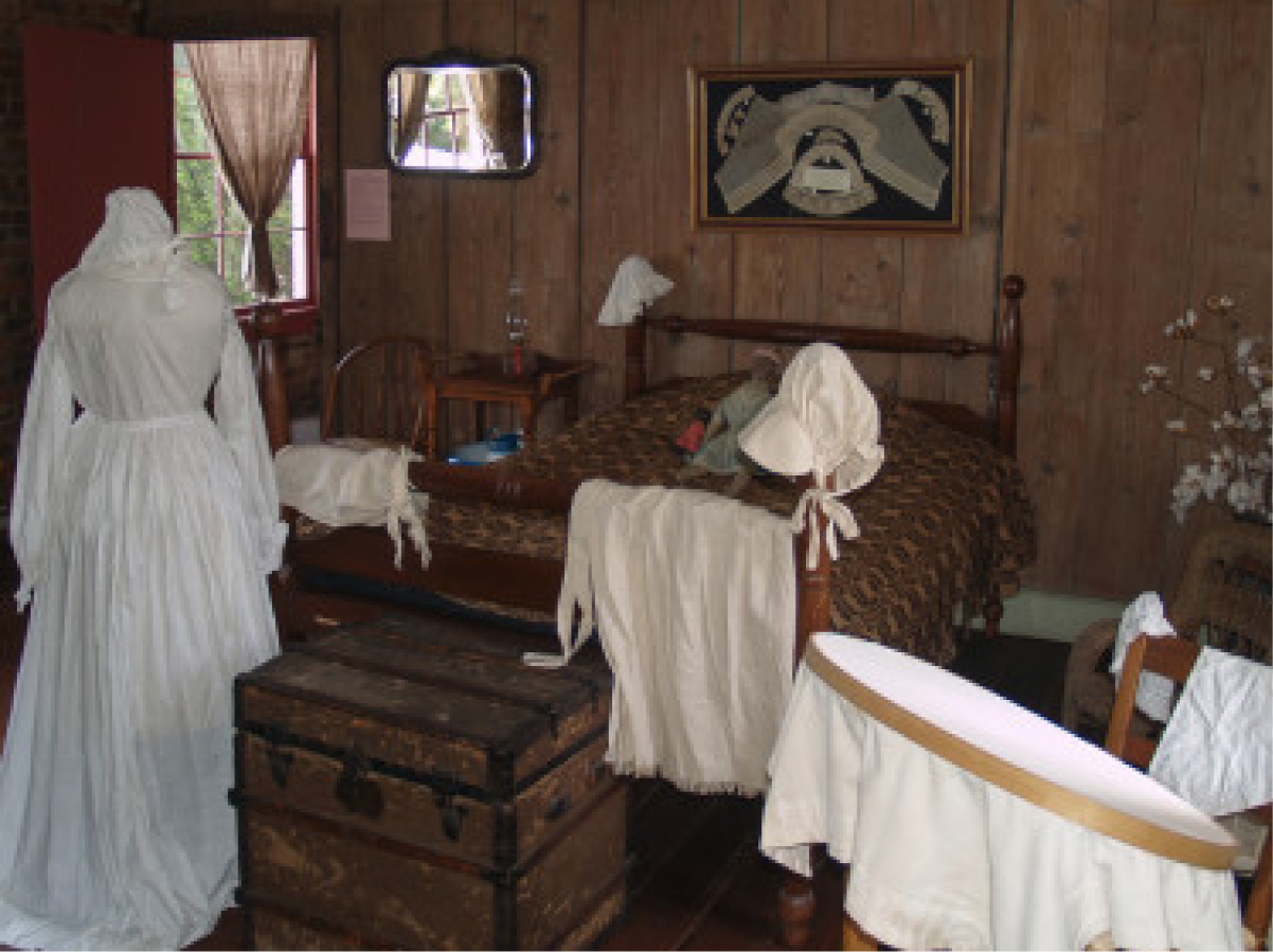 Historical bedroom with wooden walls, patterned bedding, white garments, dress form, spinning wheel, chest, and framed art above bed. Natural light enters through curtained window.