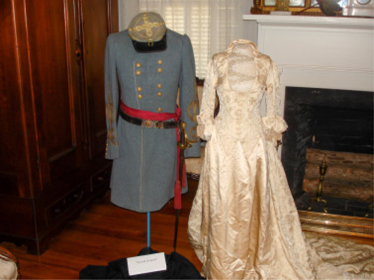 A display of two vintage outfits on mannequins, one a gray military uniform with red sash, the other a cream lace-trimmed gown.