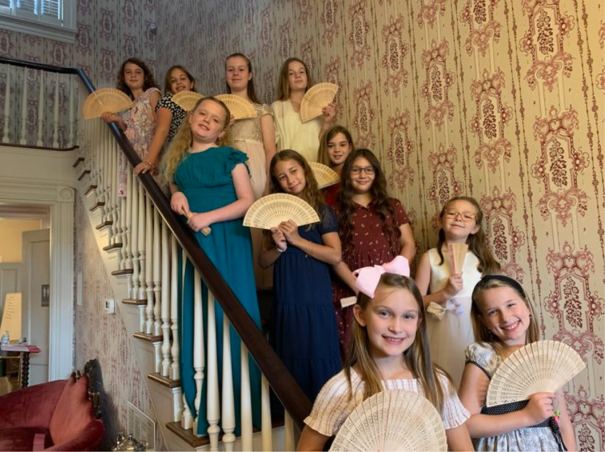Eleven girls in vintage dresses pose with hand fans on a staircase and floor, smiling in a room with patterned wallpaper.