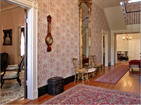 Hallway with patterned wallpaper, large ornate mirror, wooden floors, red rugs, two chairs, wall clock, and antique decor.