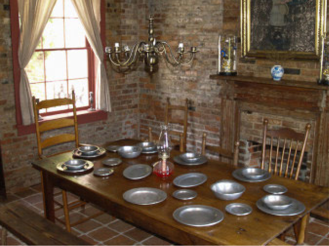 Rustic dining room with a wooden table set with metal plates, wooden chairs, brass chandelier, brick wall, window, and fireplace.