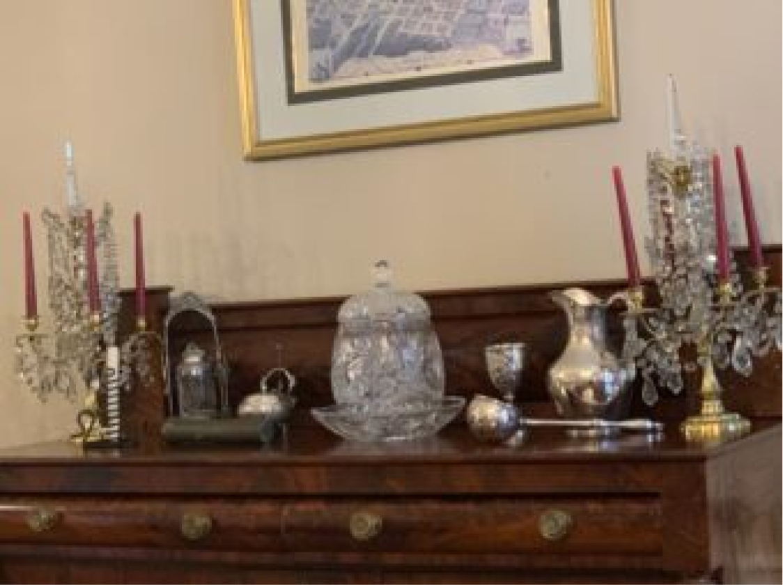 A polished wooden sideboard holds crystal candelabras with red candles, silverware, a glass dish, and framed art above on a beige wall.