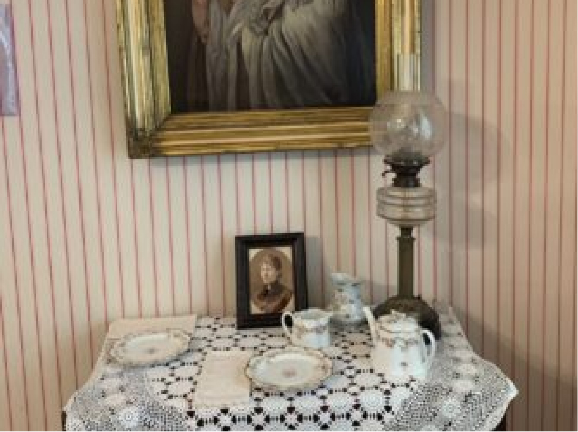 A small table with a lace tablecloth displays vintage china, a framed portrait, and an oil lamp under a large painting on striped walls.