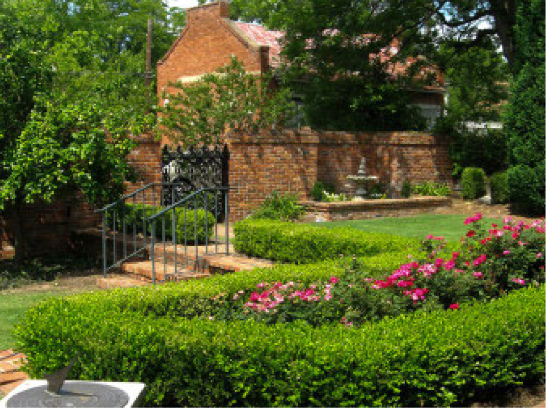 Lush garden with trimmed hedges, pink flowers in bloom, brick walkway and railing, small fountain before a brick house and trees.