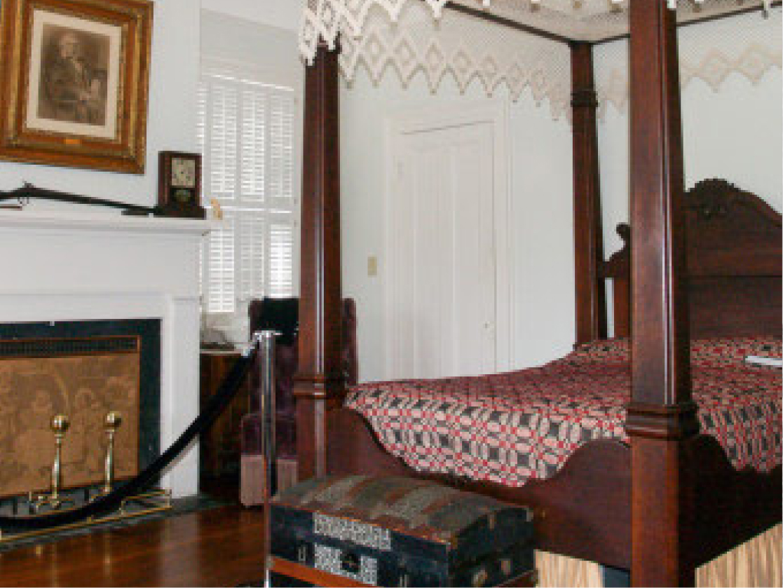A vintage bedroom with a four-poster bed, patterned quilt, white fireplace and brass tools, framed art, and sunlight through shutters.