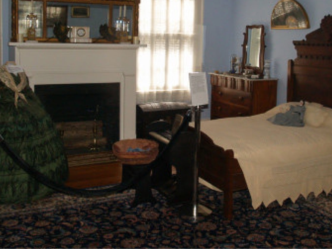 Historic bedroom with wooden bed, cream blanket, ornate dresser, fireplace, period decor, display dress, velvet rope, sunlight.