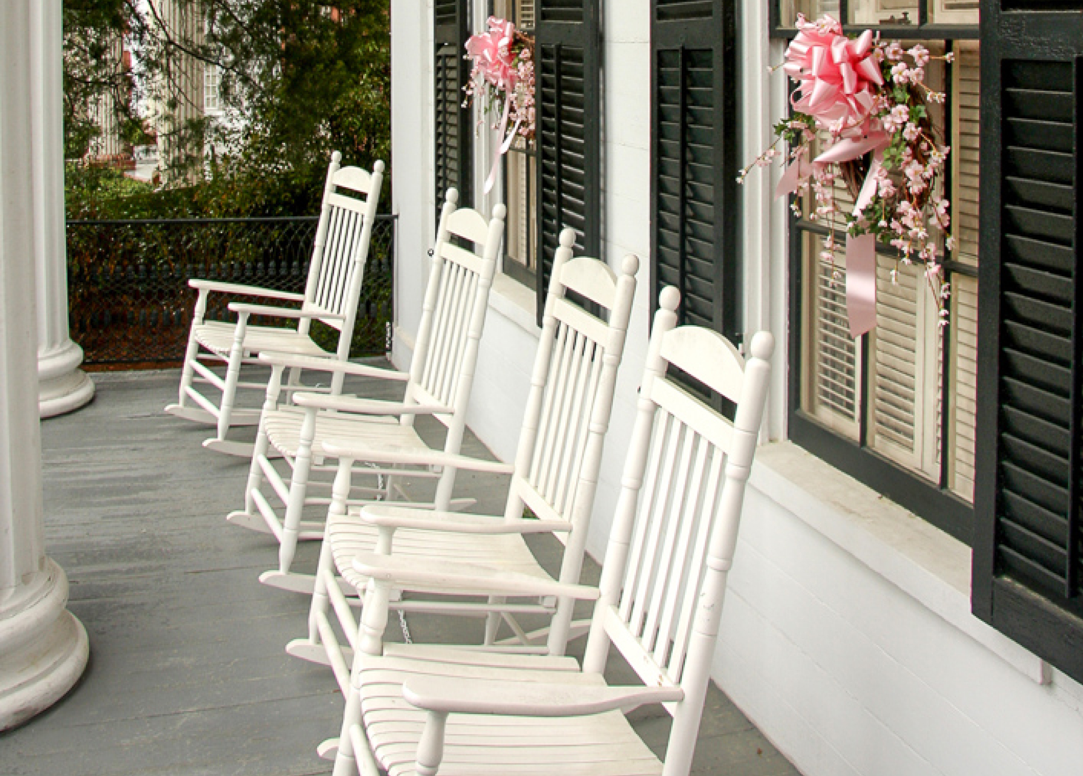 Four white rocking chairs on a porch near shuttered windows with pink ribbons and flowers, creating a peaceful southern vibe.