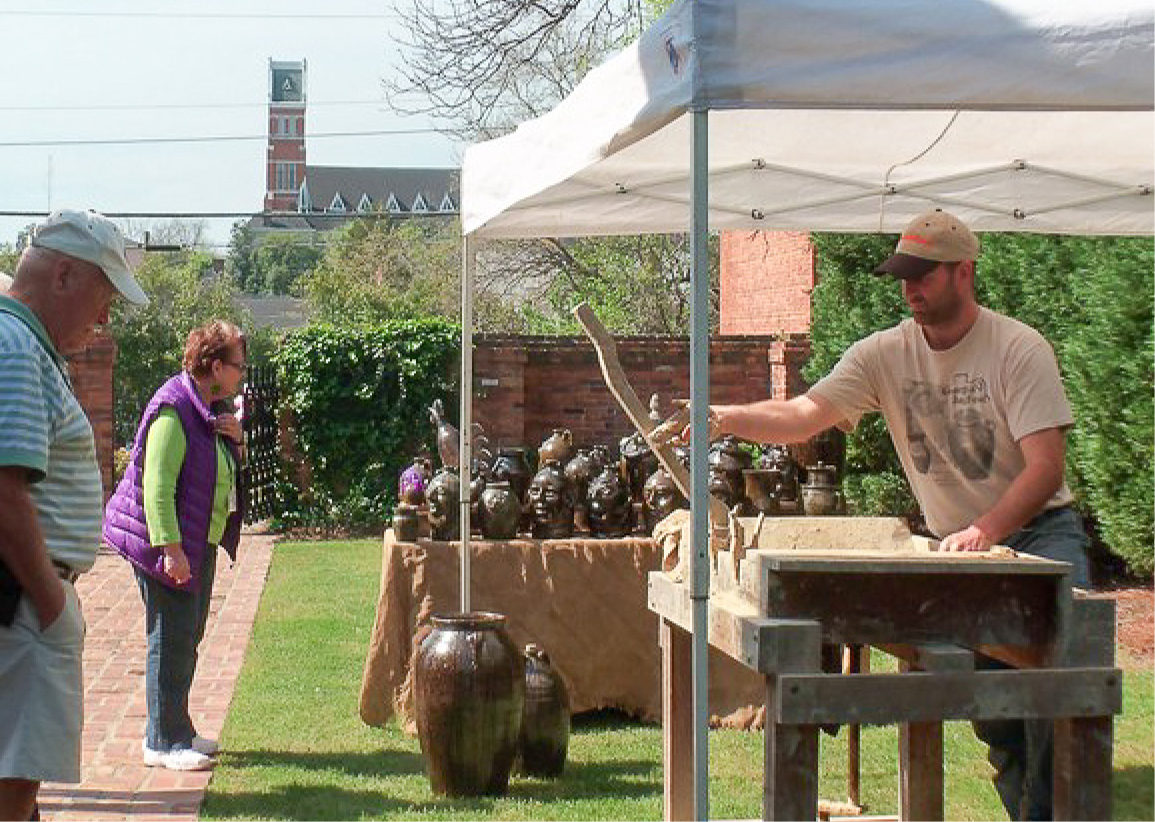 A man makes pottery under a canopy at an outdoor market, watched by a woman and older man. Pottery sits on a table; clock tower behind.