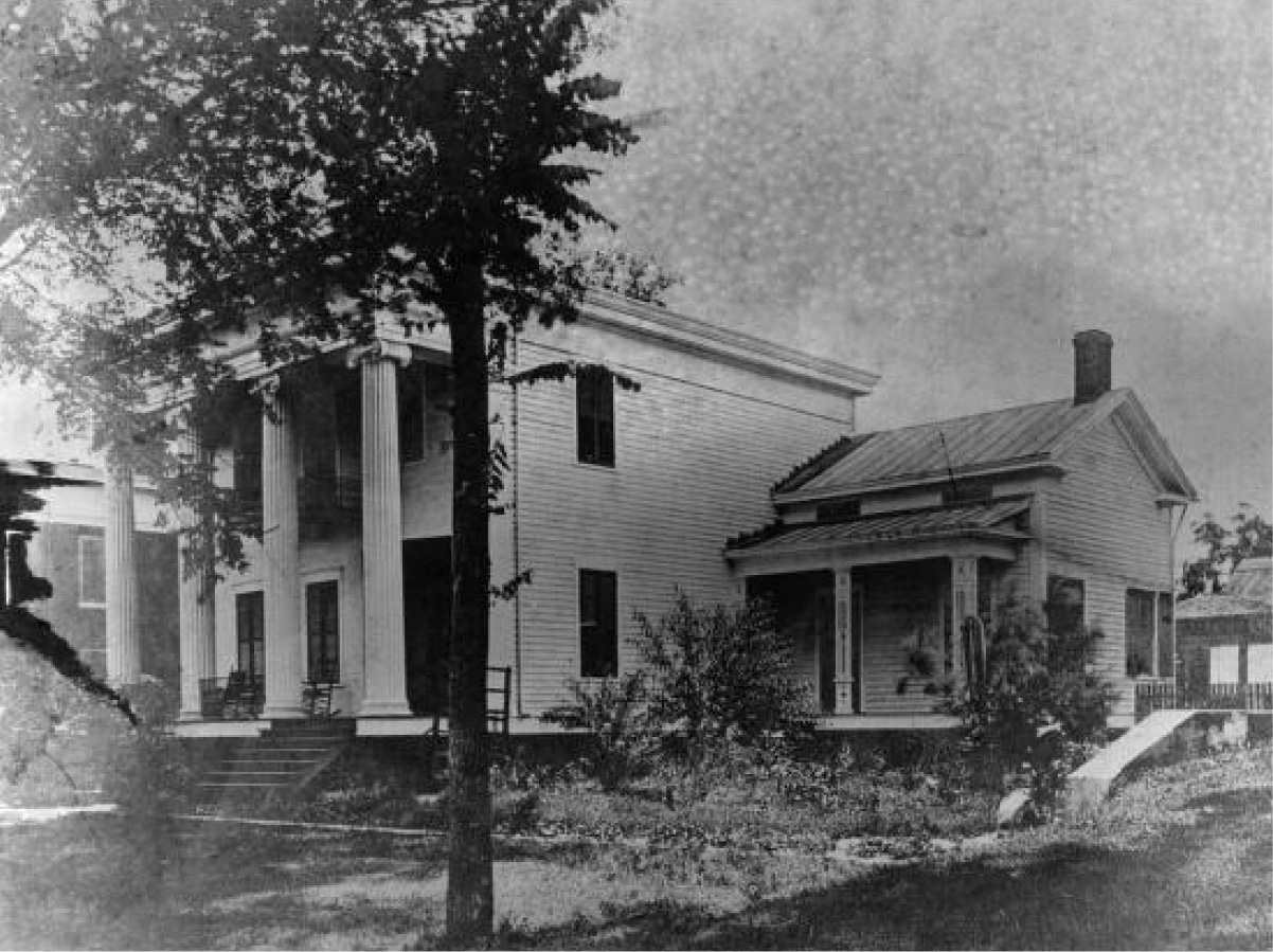 Black-and-white photo of a two-story wooden house with prominent entrance columns, side porch, and trees and shrubs in front.