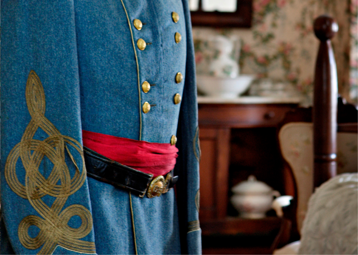 Close-up of a blue military coat with gold buttons, gold embroidery, red sash, and black belt in a room with floral wallpaper.