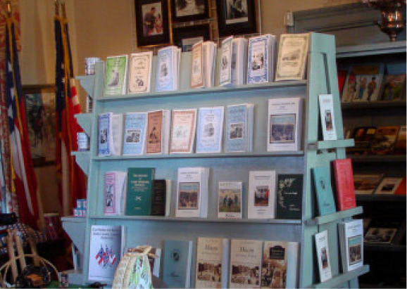 A wooden display shelf holds history pamphlets and booklets. American flags stand behind it, with framed photos on the wall nearby.