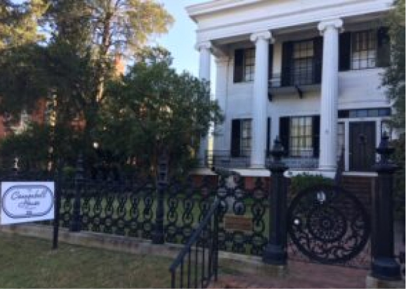 A historic white house with tall columns and black shutters sits behind a wrought-iron fence labeled Campbell House, with trees and lawn.