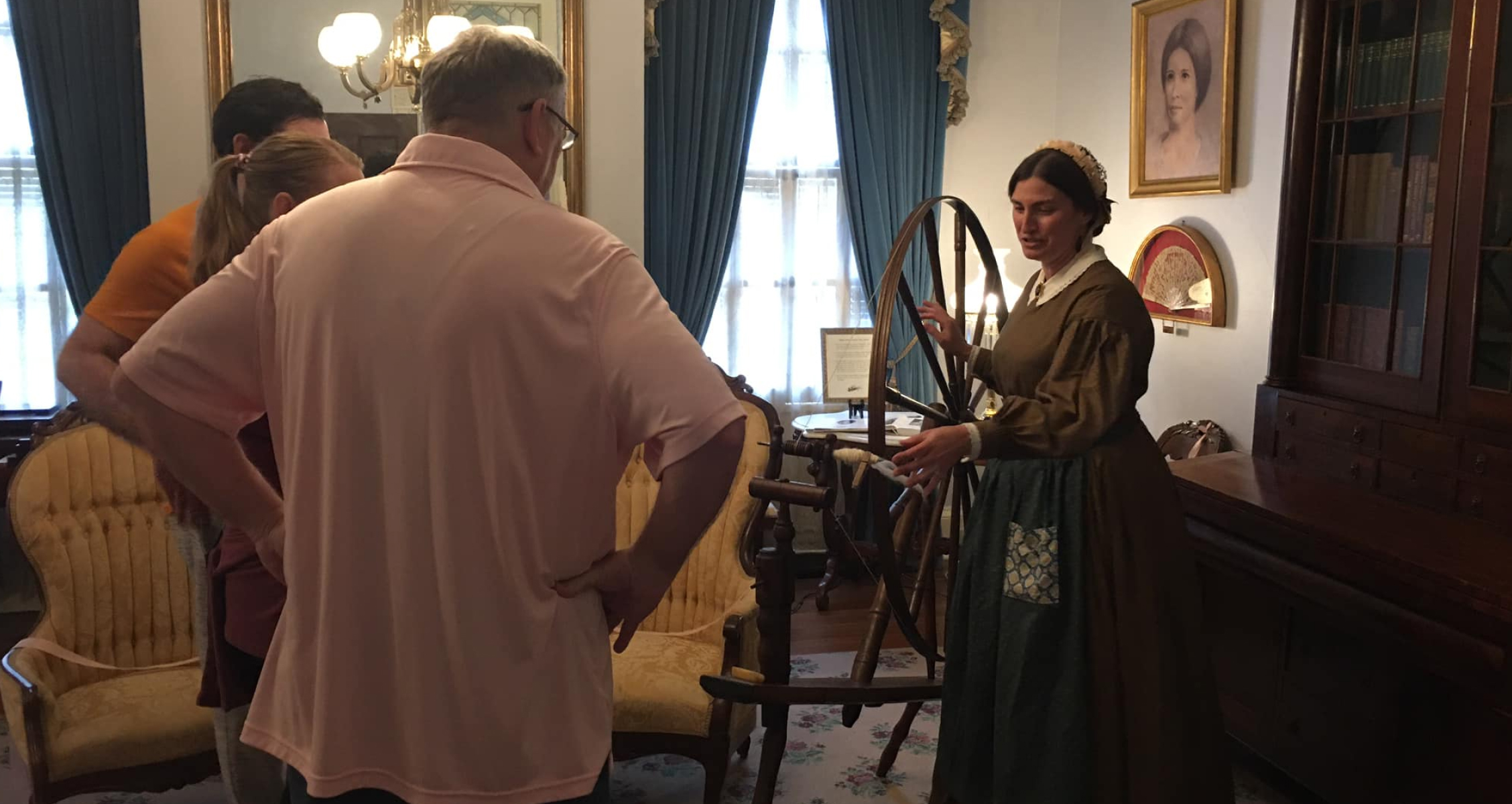 A woman in period dress uses a spinning wheel for a small audience in a Victorian parlor with ornate furniture and blue curtains.