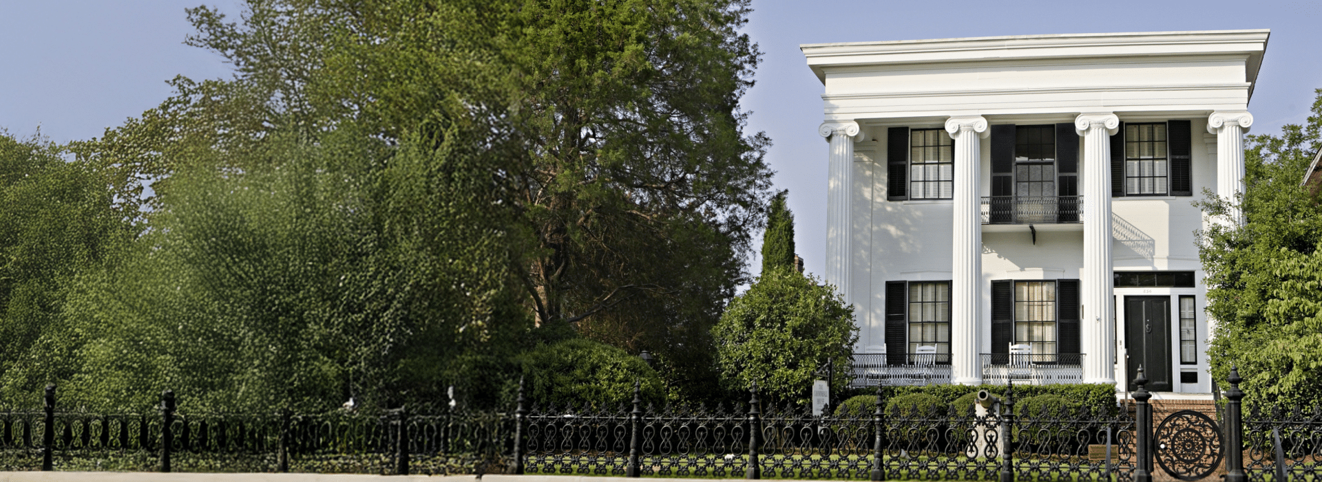 Large white house with tall columns and black trim behind a wrought iron fence, surrounded by green trees under a clear blue sky.
