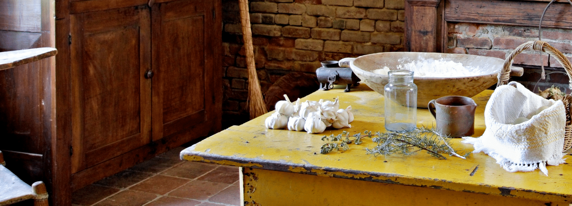 Rustic kitchen with a yellow table topped with garlic, herbs, flour, and jars, before a brick wall and wooden cabinets.
