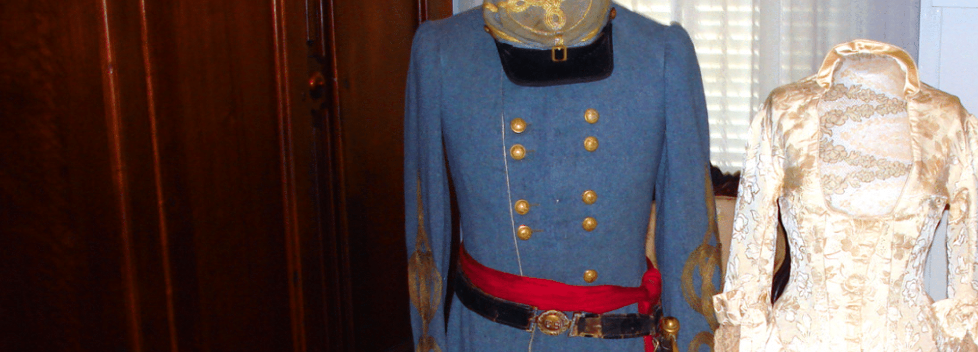 Two vintage garments on display: a blue military uniform with gold buttons and red sash, and a cream lace high-collar dress on mannequins.