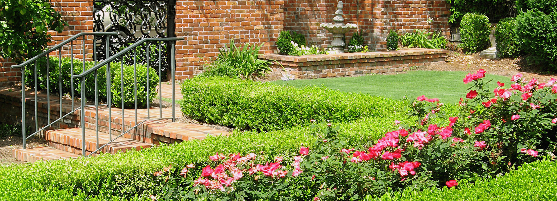 Landscaped garden featuring pink flowers, trimmed hedges, green lawn, brick path with iron railings, and a stone fountain.