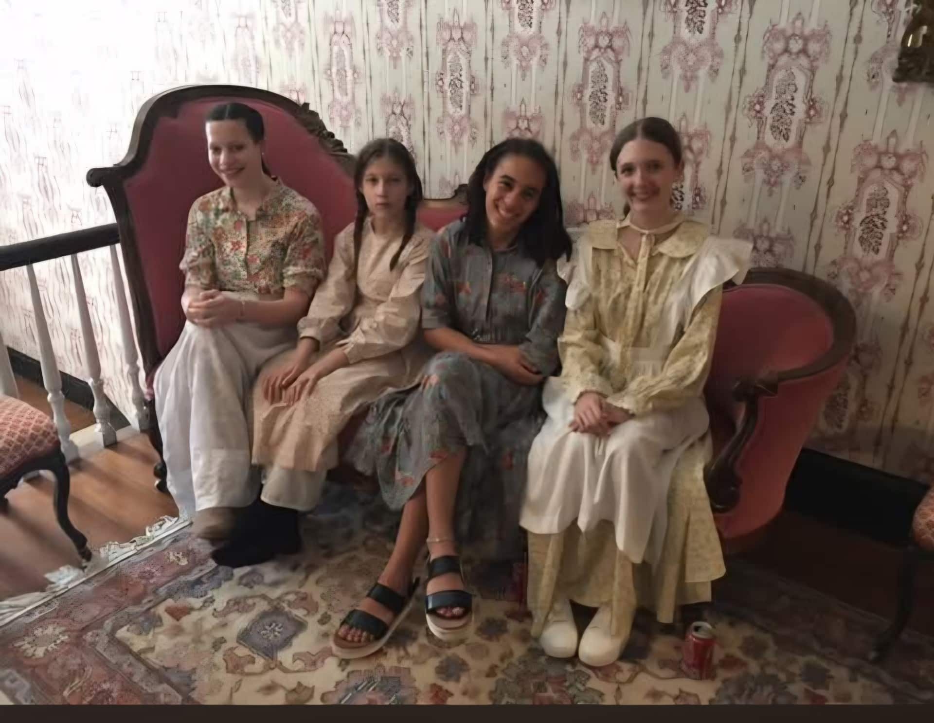 Four girls in vintage dresses are seated on a pink antique sofa in a room with floral wallpaper. One of the girls stands out in modern sandals paired with a floral dress. A soda can rests on the floor near their feet.